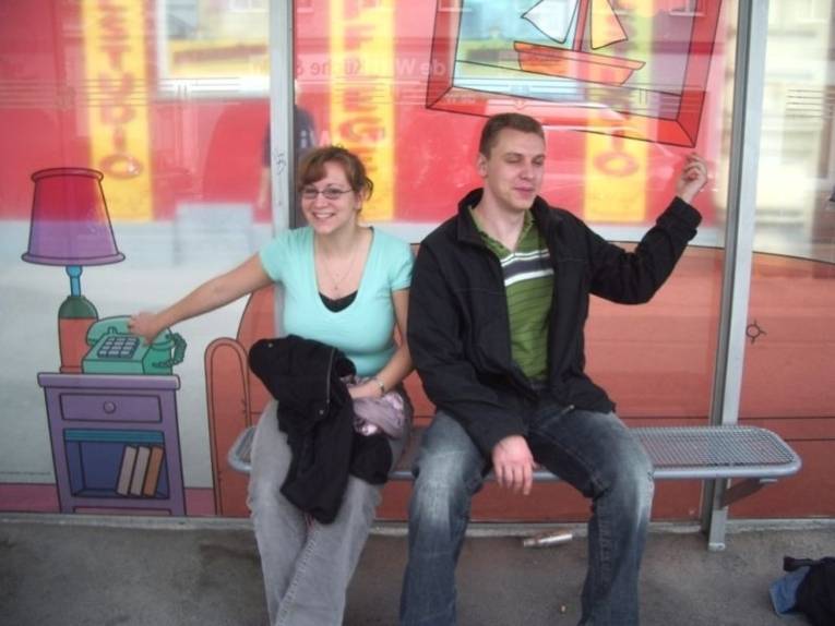 Farbfoto.Man sieht ein junges Paar sitzend auf einer Bank in einer Kulisse.